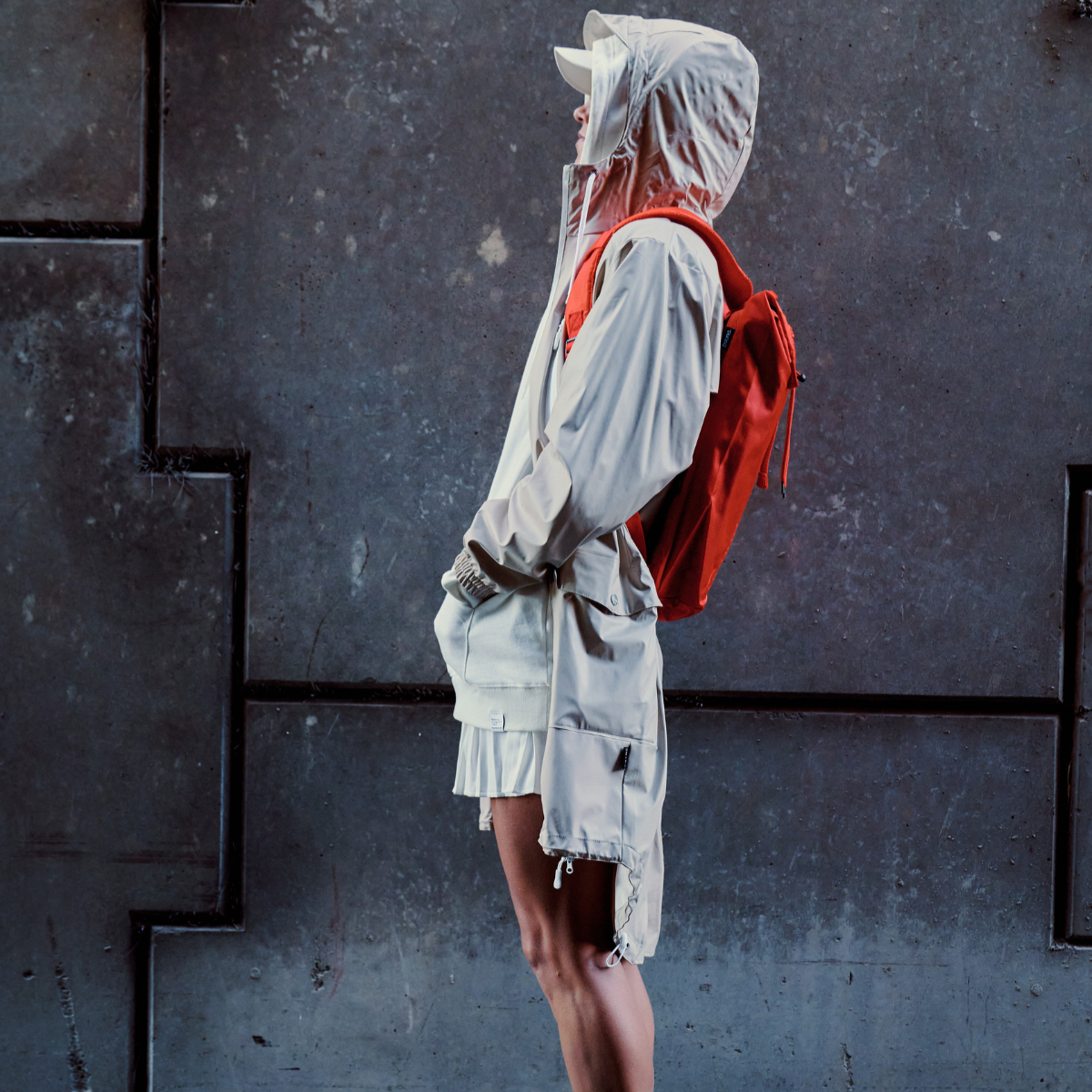 Person wearing a light-colored jacket and shorts with a red backpack against a dark wall.
